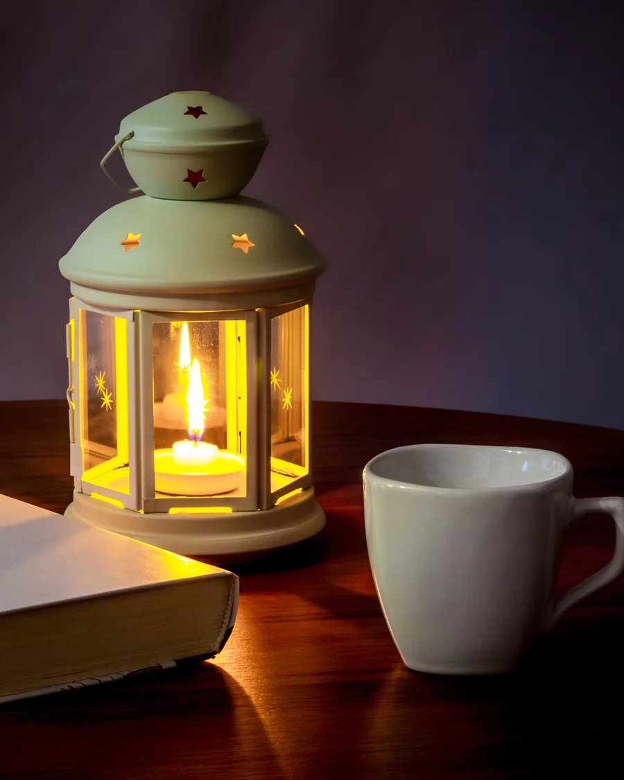 A candle lantern, a closed book, and a coffee cup on a dark table